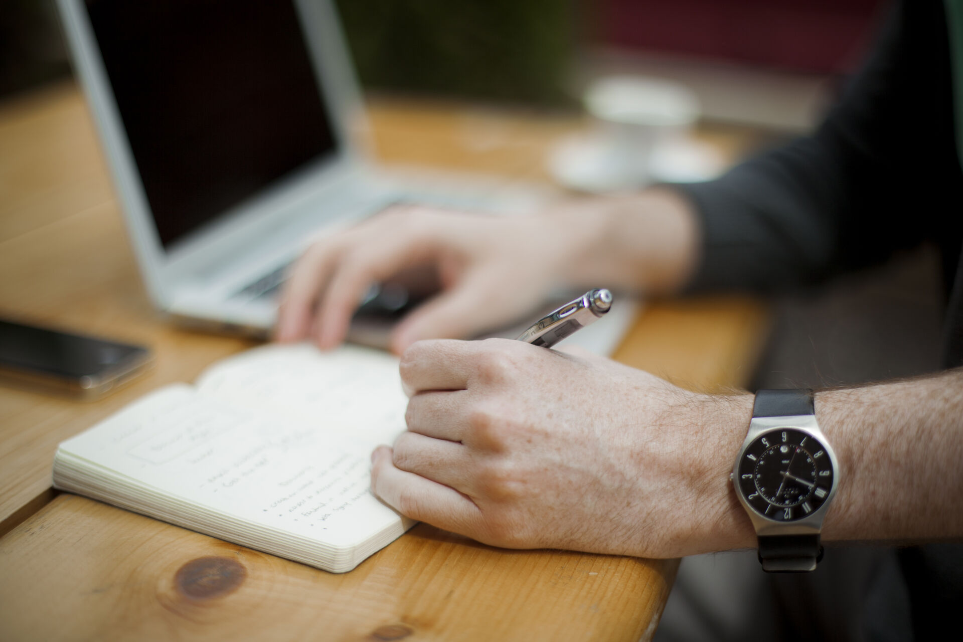 Hand writing notes in a notebook beside a laptop